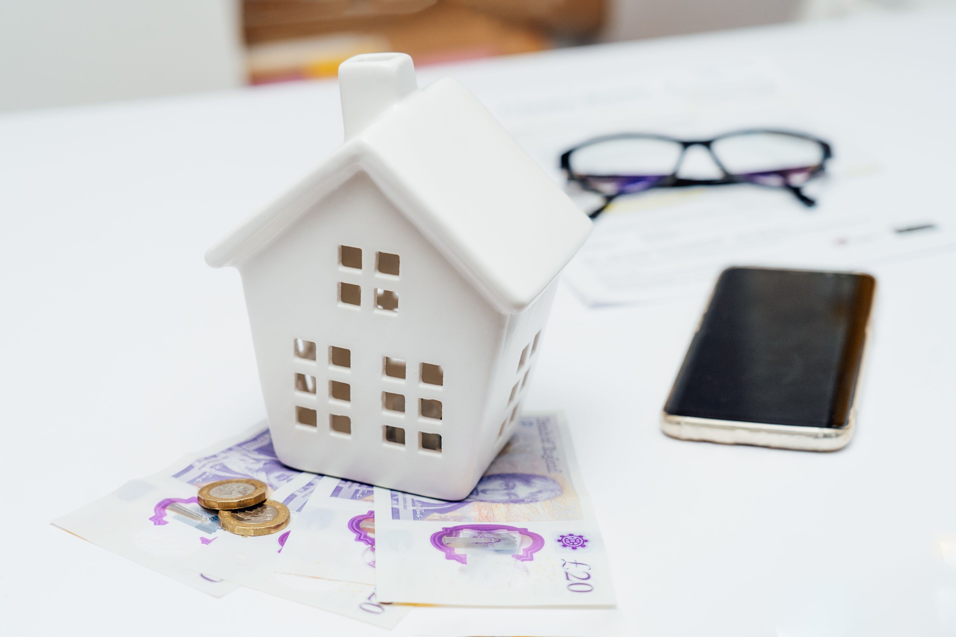 House model with British currency, bill documents and phone on table. Housing cost and maintenance payments. Real estate or property investment. Home mortgage loan rate. Saving money for retirement.